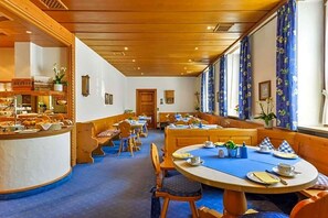 Espaço de refeições de hotel com mesas de madeira, toalhas de mesa azuis e cozinha visível ao fundo.