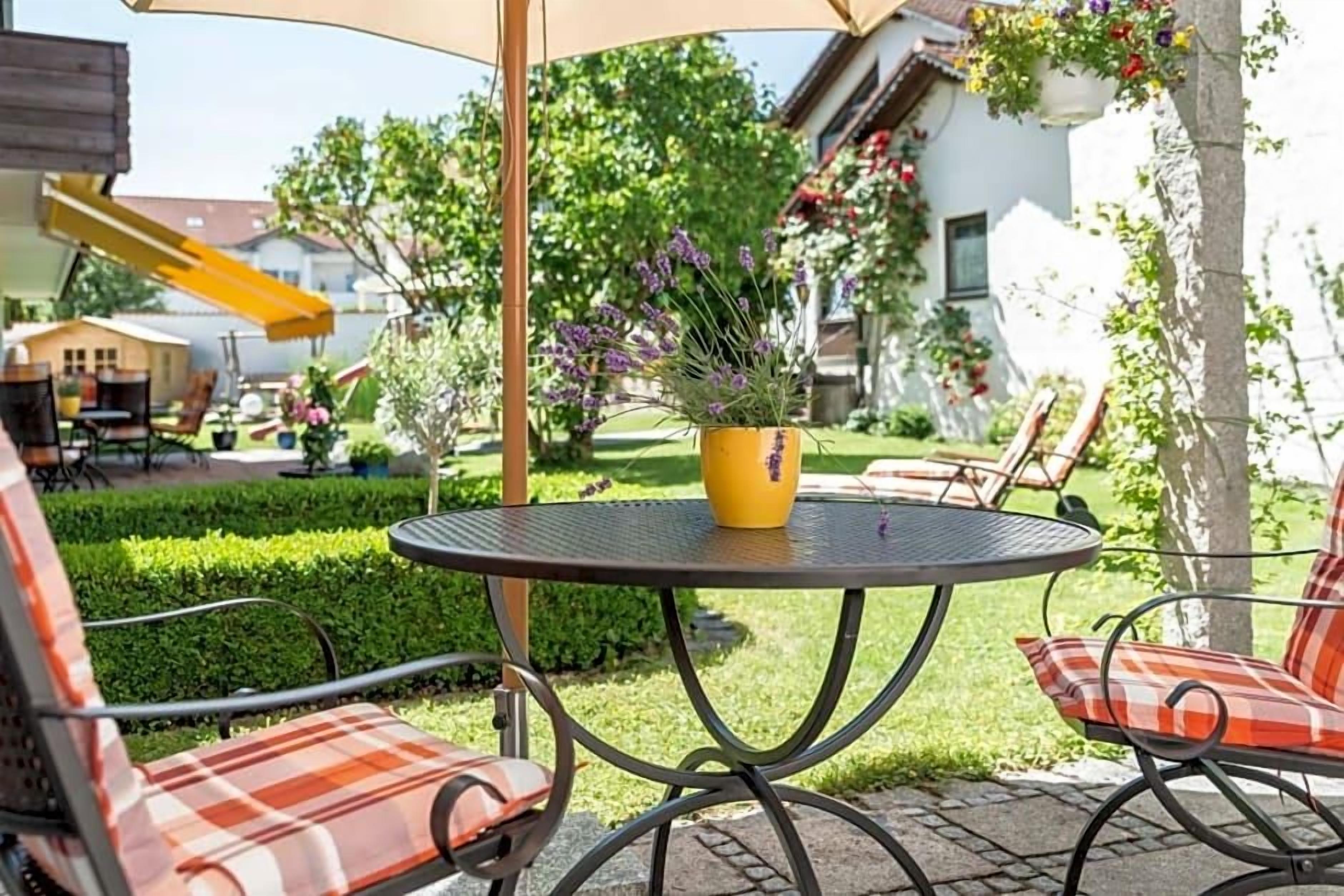 An outdoor patio with a round table and chairs, a yellow planter with flowers, and a yellow slide in the background.
