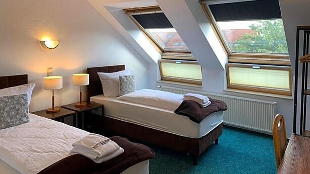 Quarto de hotel com duas camas, uma janela, um radiador e uma mesa de cabeceira com luminárias.