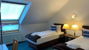Quarto de hotel com teto inclinado, claraboia, duas camas, abajures, escrivaninha e uma cadeira.