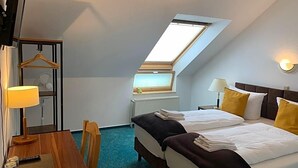 Quarto de hotel com cama, secretária, cadeira, televisão, janela e iluminação no teto.