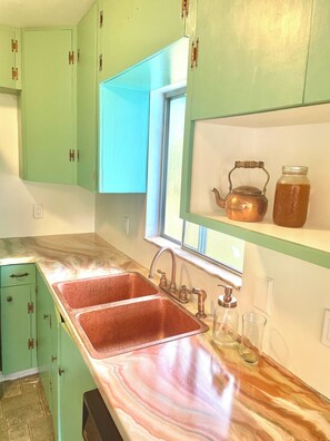 Une cuisine avec des meubles de couleur verte, un évier en cuivre et une fenêtre au-dessus du plan de travail.