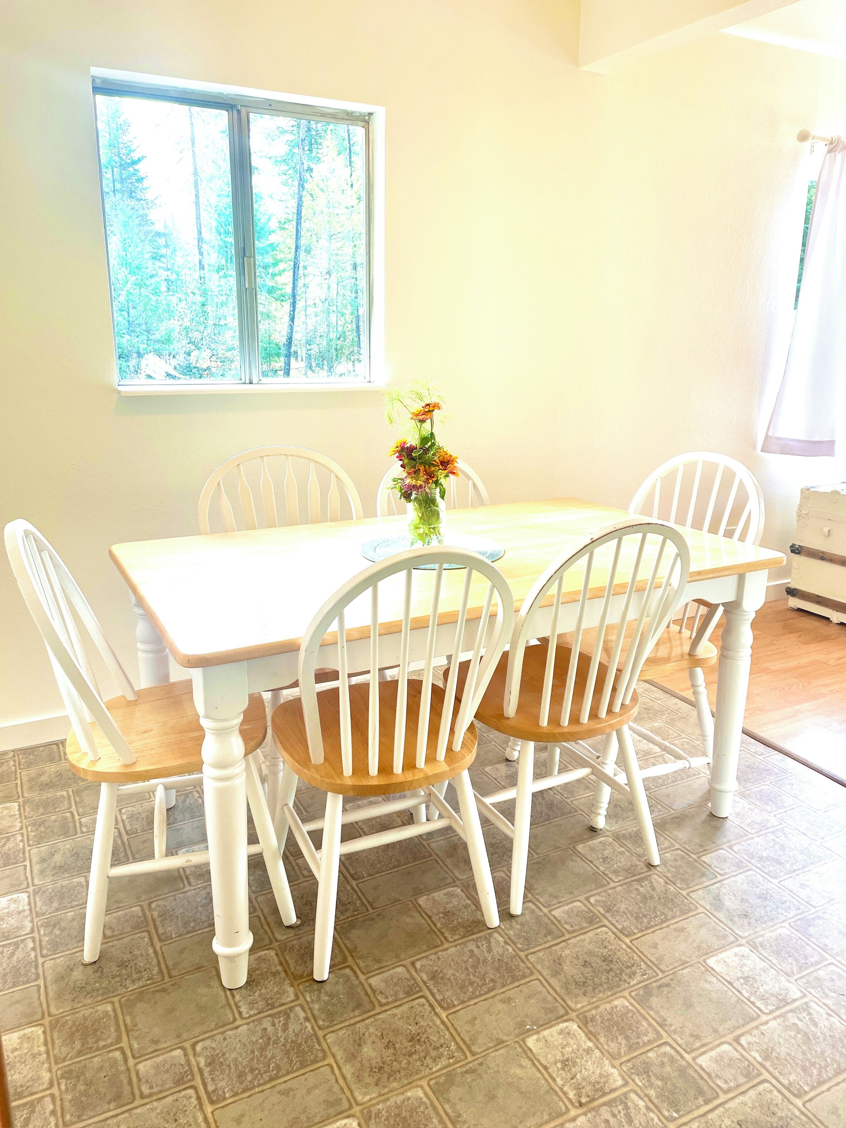 A dining area with a table and chairs, a window with a view of trees, and a vase with flowers on the table.