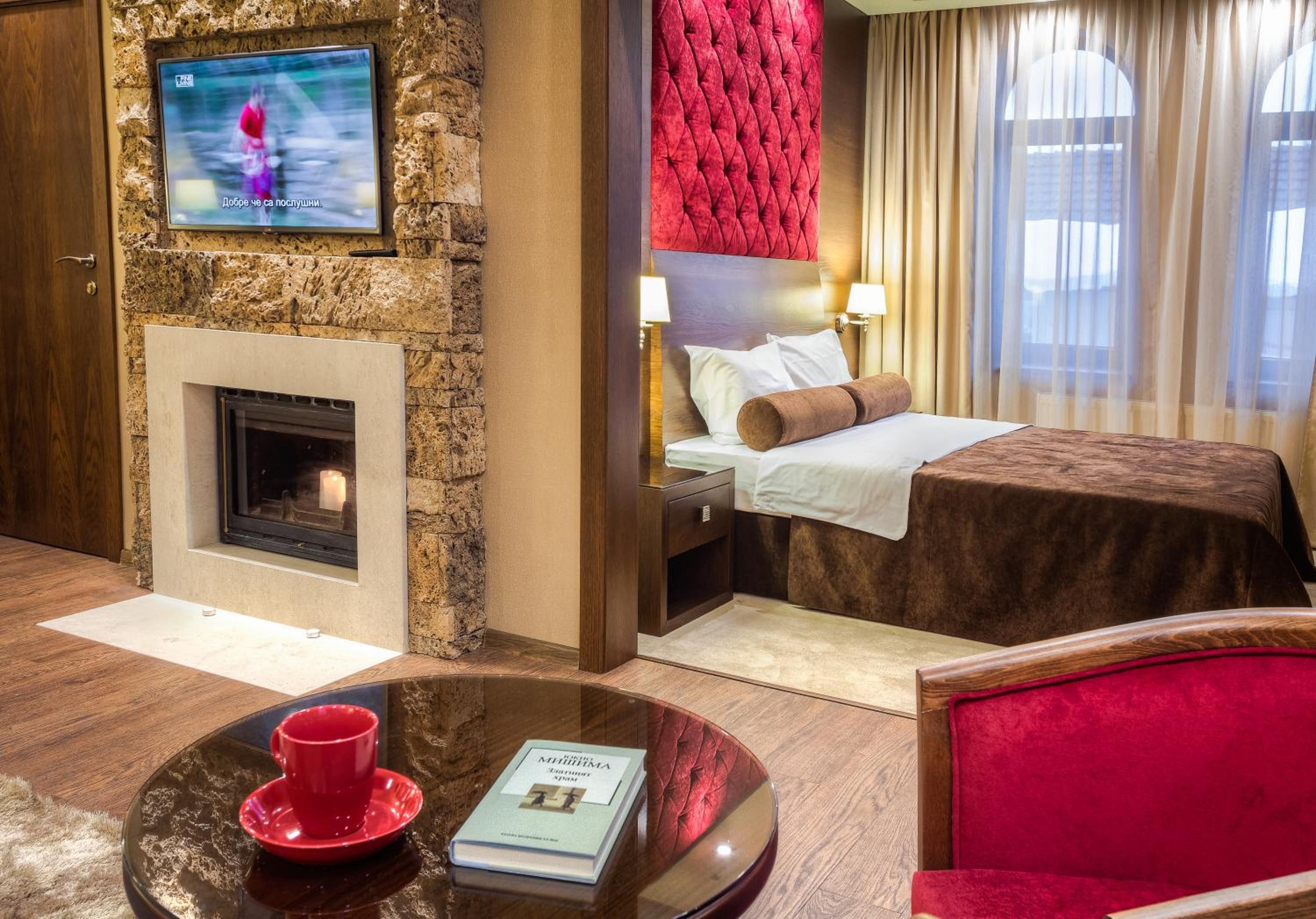 Habitación de hotel con pared de piedra, chimenea, cama con cabecero rojo, mesita de noche con lámpara y ventana con cortinas.