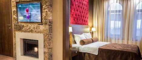 A hotel room with a stone wall, a fireplace, a bed with a red headboard, a bedside table with a lamp, and a window with curtains.
