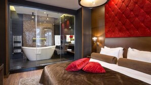 Moderni hotellihuone, jossa on suuri sänky, punainen tehosteseinä, lasinen suihkukaappi ja näkymä kylpyhuoneeseen.