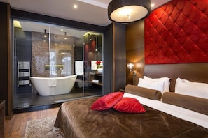 A modern hotel room with a large bed, a red accent wall, a glass shower enclosure, and a view of the bathroom. - Family Hotel Strannopriemnitza (Troyan)