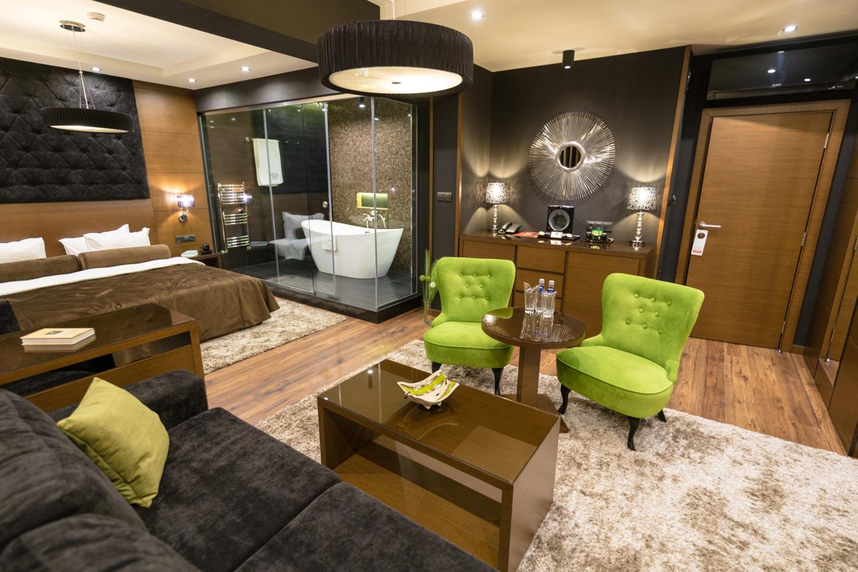 A modern hotel room with a large bed, a sitting area with a coffee table, two armchairs, a glass-enclosed bathroom, and a large mirror on the wall.