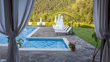 A pool area with lounge chairs and umbrellas, surrounded by a stone deck and a view of a forested hillside.