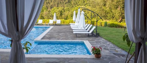 Un área de piscina con sillas de descanso y sombrillas, rodeada por un solárium de piedra y vistas a una ladera boscosa.