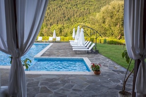 A pool area with lounge chairs and umbrellas, surrounded by a stone deck and a view of a forested hillside. - Family Hotel Strannopriemnitza (Troyan)
