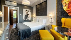 Moderni hotellihuone, jossa on sänky, yöpöydät, keltainen nojatuoli ja suuri abstrakti maalaus.