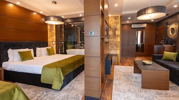 A modern hotel room with a large bed, a sitting area with a coffee table, and a separate living area with a sofa and a TV.