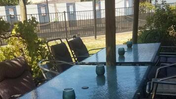 An outdoor covered dining area with a table and chairs, surrounded by foliage and a fence.