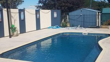 A rectangular swimming pool with a concrete deck, surrounded by a metal fence and a building with a green roof.