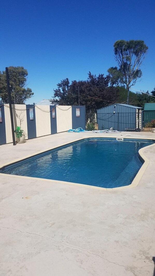 A rectangular swimming pool with a concrete deck, surrounded by a metal fence and a building with a green roof. - Wimmera Motel (Nhill)