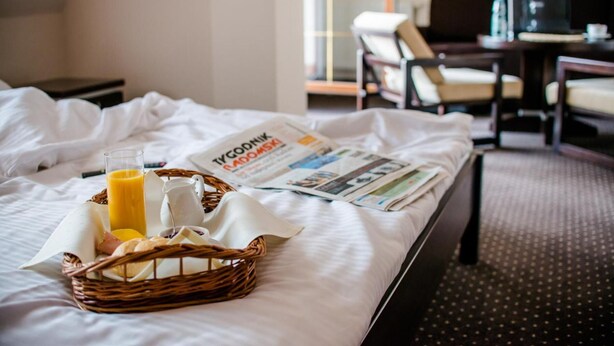 Habitación de hotel con una cama, una bandeja de desayuno con pan, un vaso de zumo de naranja, un periódico y una cesta de mimbre.