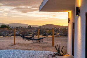 A desert landscape with a hammock between wooden posts, a building with a modern design, and a clear sky. - Hōkū Hale | Spa | Sauna | Fire Pit | 360° Views (Joshua Tree)