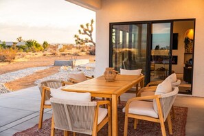 Une terrasse extérieure avec une table en bois et des chaises en osier, une porte vitrée donnant sur un espace intérieur, et un paysage désertique en arrière-plan.