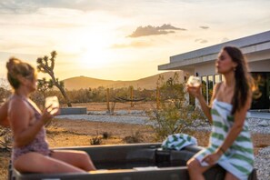 Two individuals seated in a vehicle with a desert landscape and a modern building in the background. - Hōkū Hale | Spa | Sauna | Fire Pit | 360° Views (Joshua Tree)