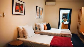 A hotel room with two beds, a small table, a red blanket, and a door leading to a bathroom.