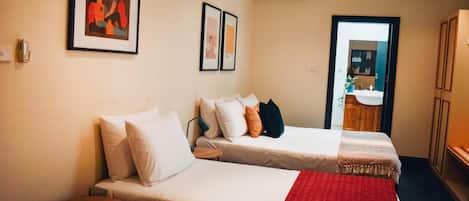 Une chambre d’hôtel avec deux lits, une petite table, une couverture rouge et une porte donnant sur une salle de bain.