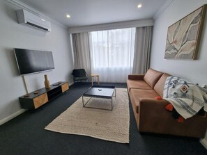 A modern living room with a flat-screen TV, a brown sofa, a coffee table, and a large window with curtains.