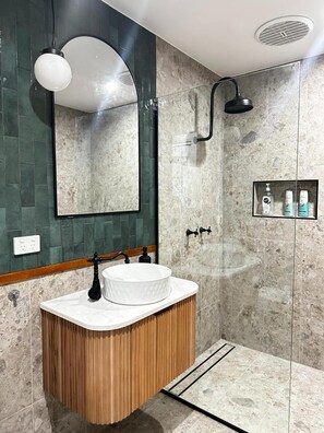 A bathroom with a round sink, a mirror, a shower, and a shelf with toiletries.