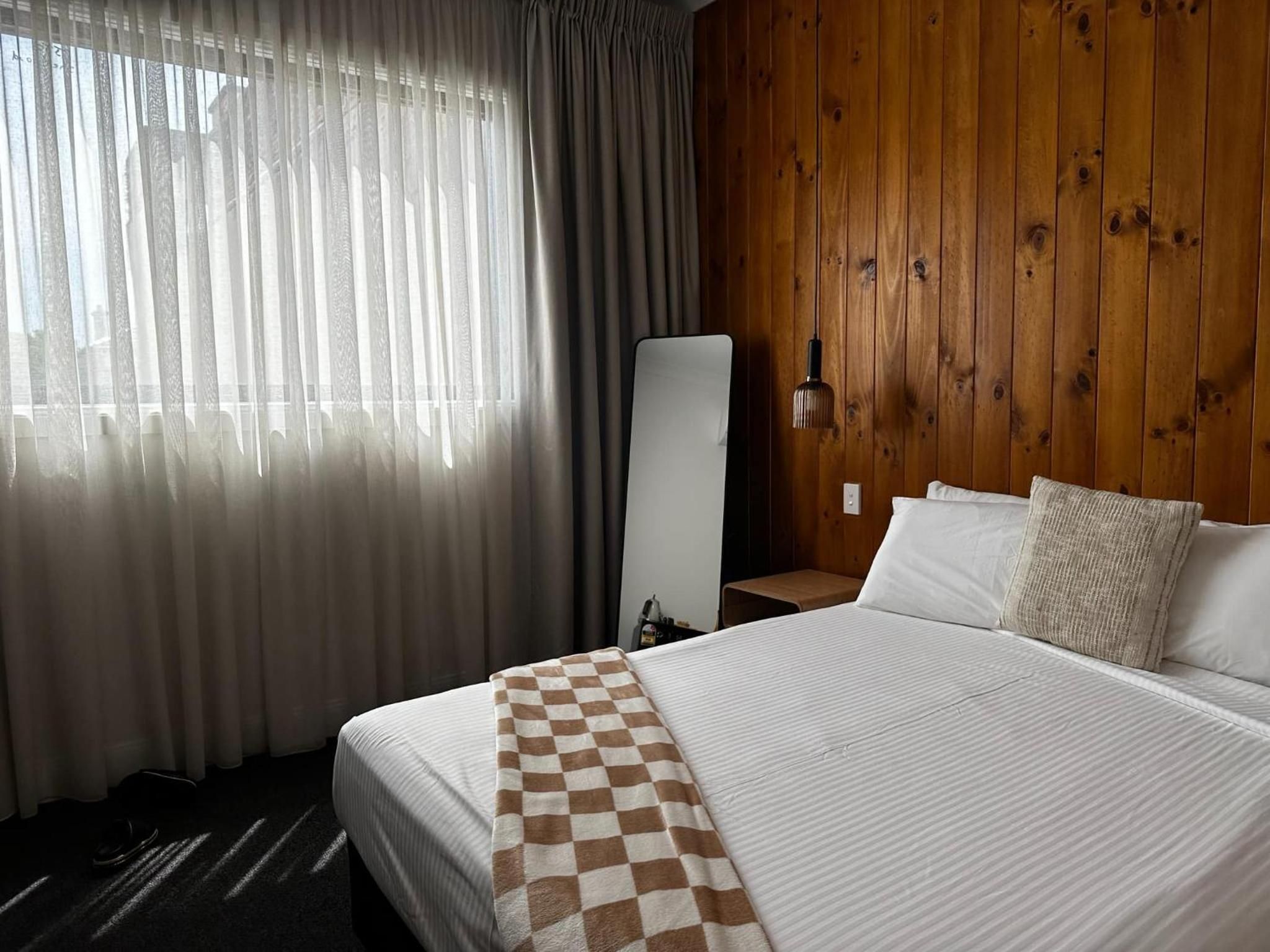 A hotel room with a large bed, a checkered throw blanket, a wooden wall, and a window with sheer curtains.