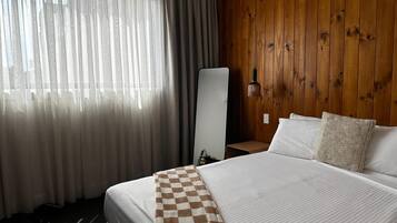 A hotel room with a large bed, a checkered throw blanket, a wooden wall, and a window with sheer curtains.