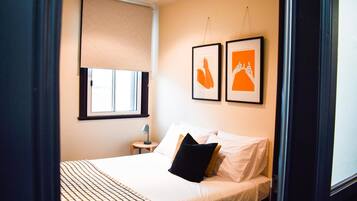 Une chambre à coucher comprenant un lit, une table de chevet, une lampe, une fenêtre avec un store et deux tableaux encadrés accrochés au mur.