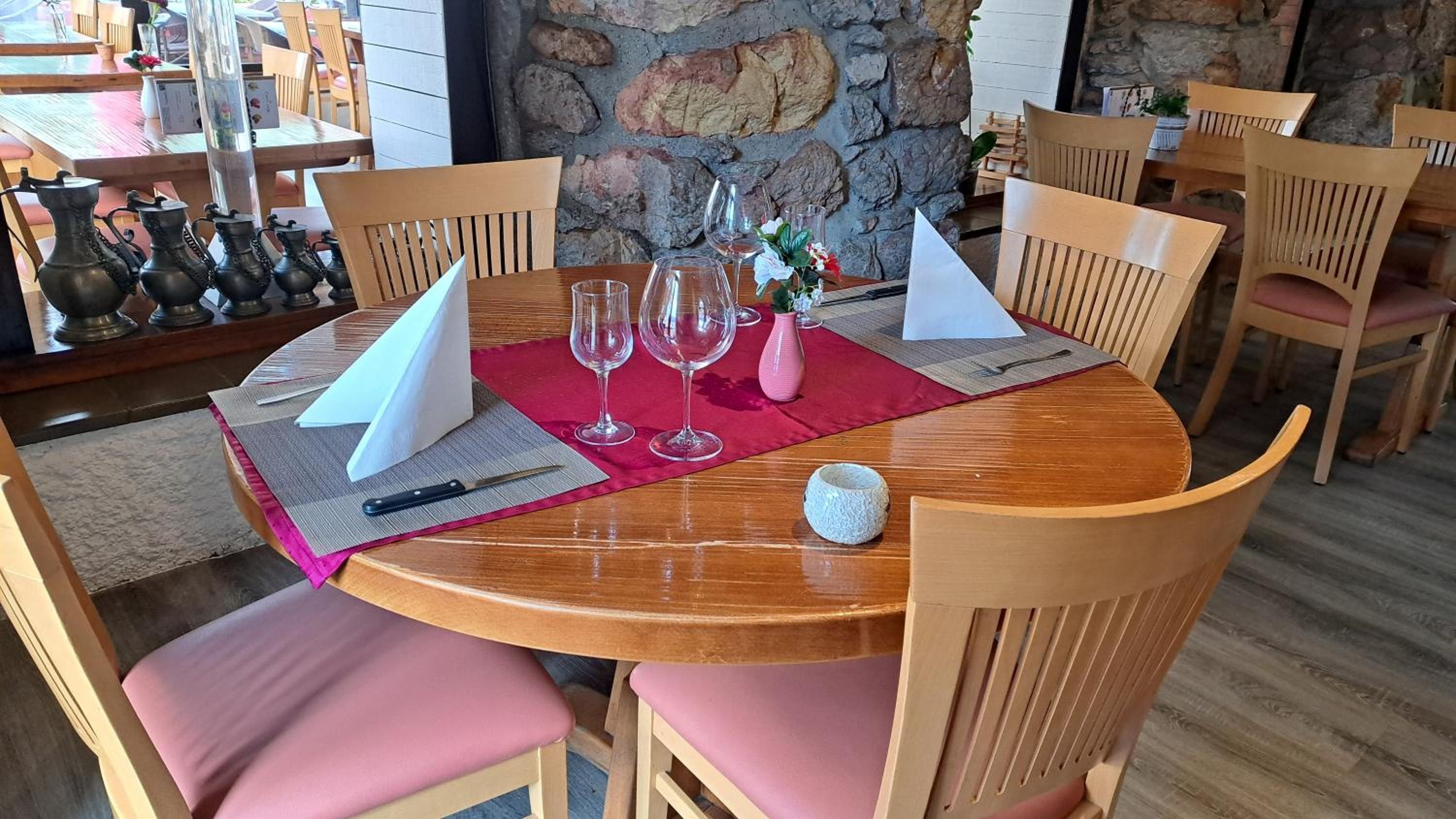 A round wooden dining table with pink cushions, set with wine glasses, a knife, and a small vase.