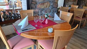 A round wooden dining table with pink cushions, set with wine glasses, a knife, and a small vase.