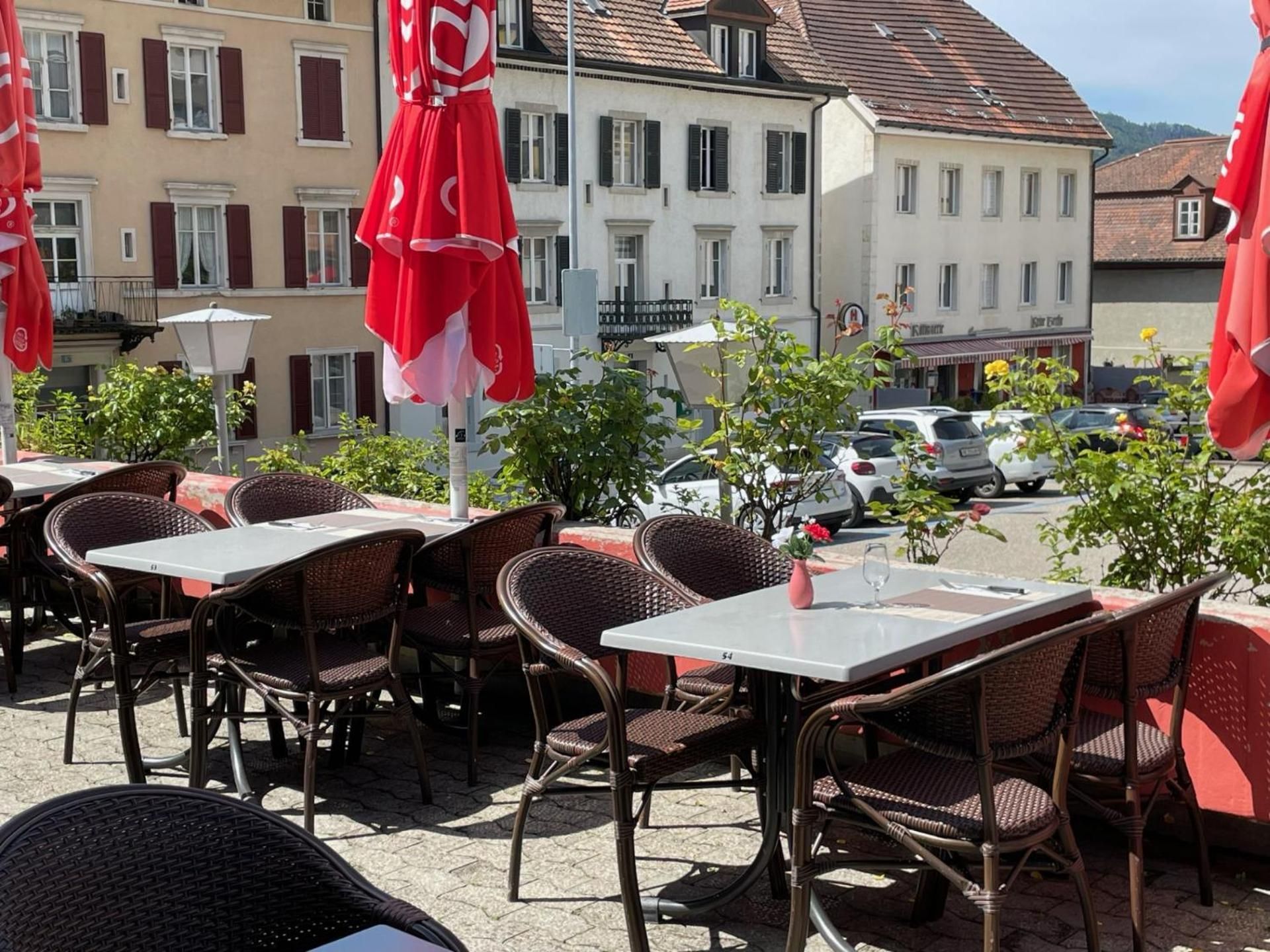 Outdoor seating with wicker chairs and tables, red umbrellas, and a street view with buildings and parked cars.