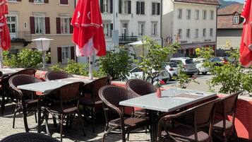 Outdoor seating with wicker chairs and tables, red umbrellas, and a street view with buildings and parked cars.