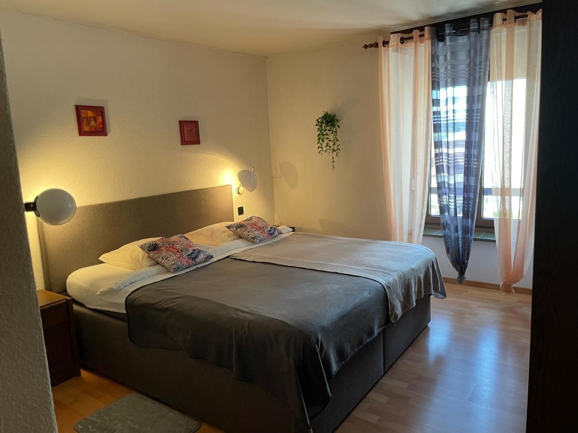 A bedroom with a bed, bedside lamps, a window with curtains, and a small plant.