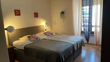 A bedroom with a bed, bedside lamps, a window with curtains, and a small plant.