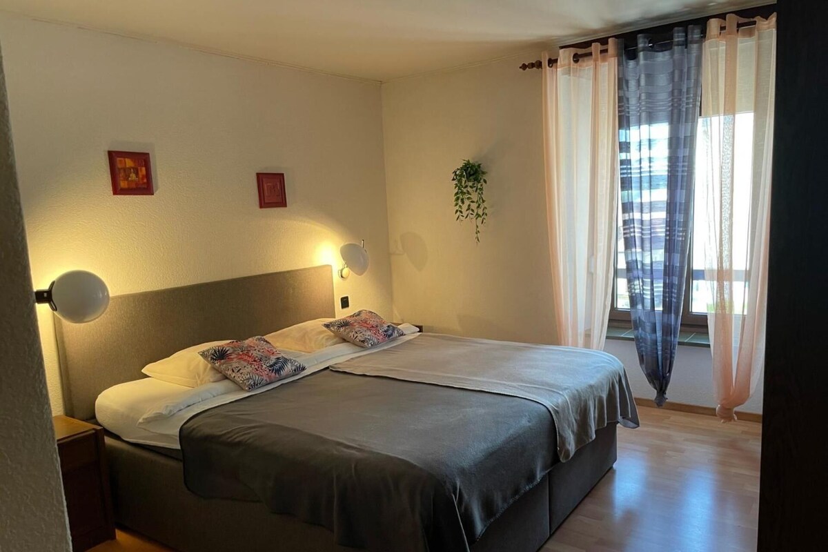 A bedroom with a bed, bedside lamps, a window with curtains, and a small plant.