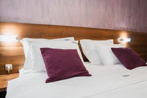 A hotel room with a wooden headboard, white bedding, and two purple pillows. - Hotel Damis (East Sarajevo)