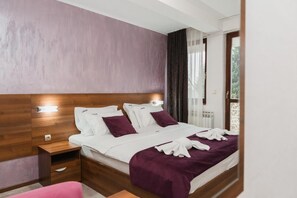 A hotel room with a large bed, wooden headboard, a nightstand, and a view of the outdoors. - Hotel Damis (East Sarajevo)