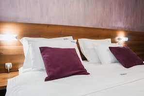 Una habitación de hotel con cabecera de madera, ropa de cama blanca y dos almohadas moradas.