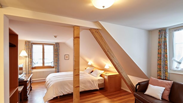 A bedroom with a sloped ceiling, wooden beams, a bed, a nightstand, a dresser, a red rug, and a brown leather sofa. - Landgasthof Hotel Rv Ssli (Adligenswil)