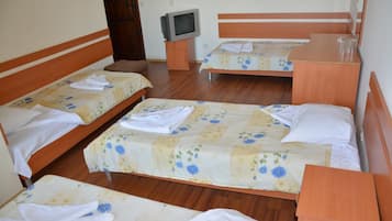 A hotel room with four single beds, each with a floral bedspread and white towels. There is a wooden nightstand with a television between the beds.