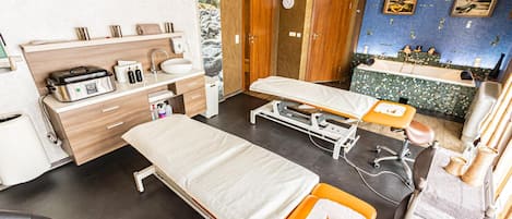 Sala de exames médicos com duas camas, pia, relógio e parede azul revestida com azulejos e obras de arte.