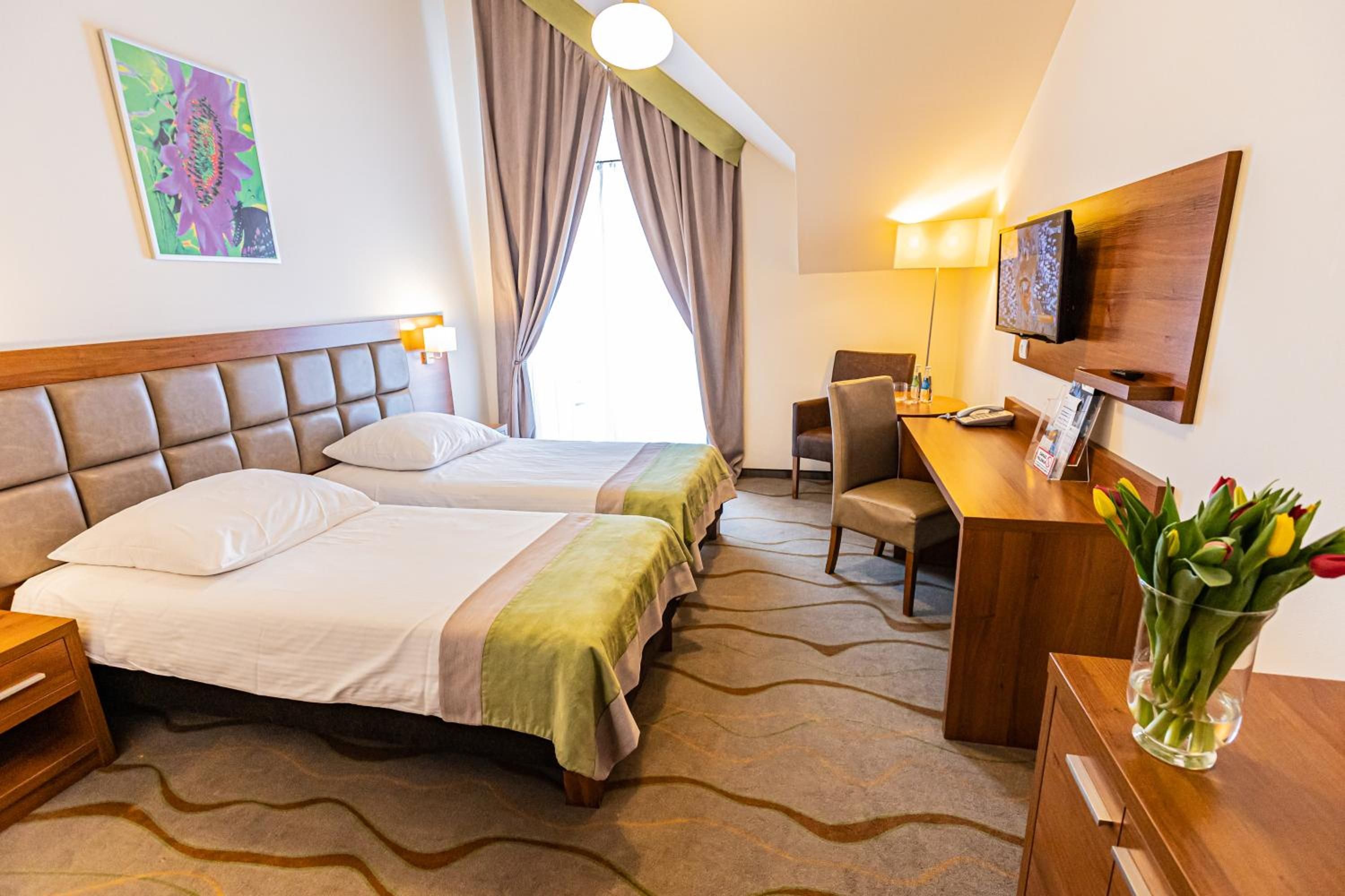 A hotel room with two beds, a desk, a chair, a TV, and a vase with flowers.