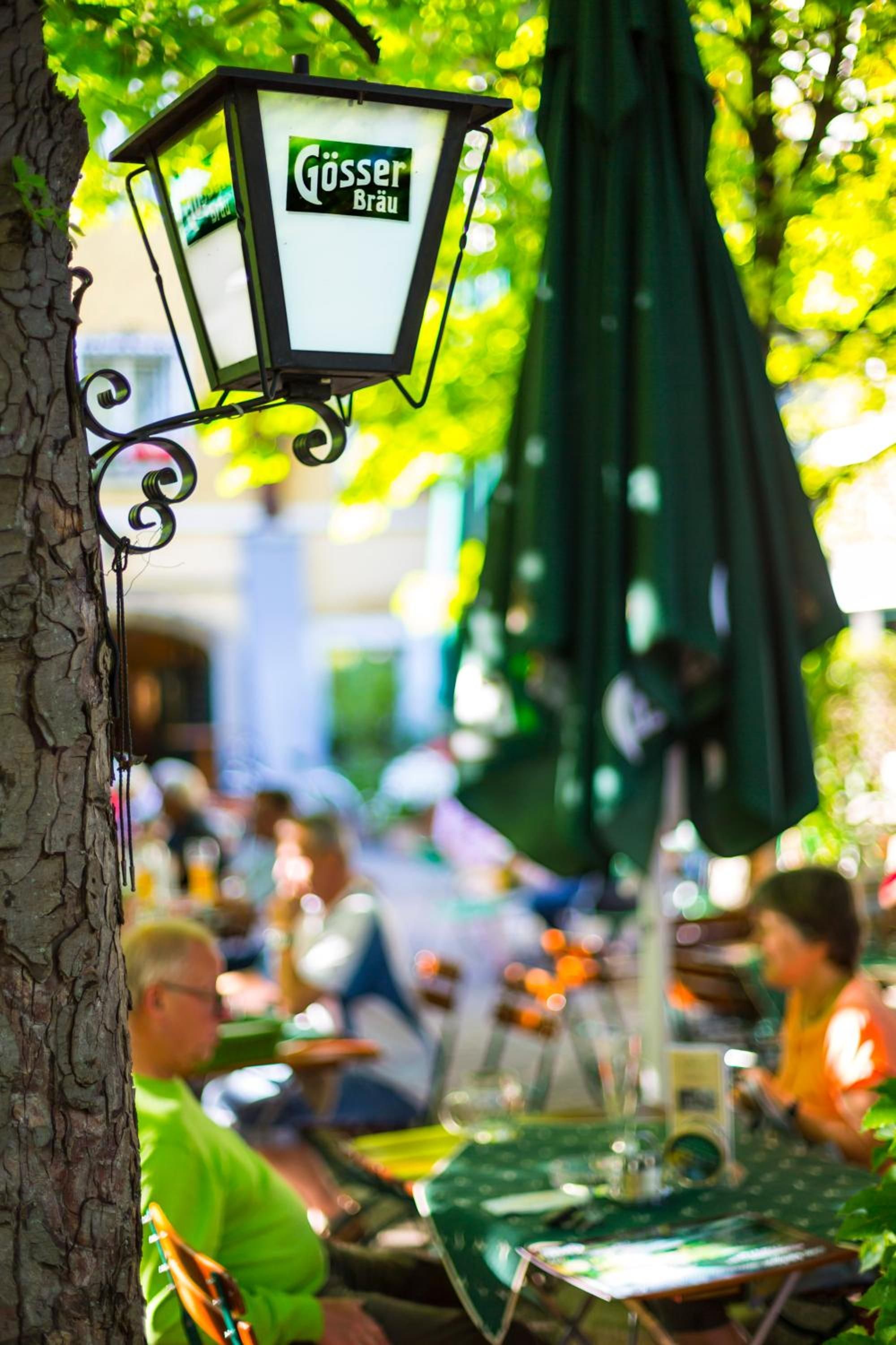 Una lámpara colgante para exteriores con un cartel, un tronco de árbol y un fondo borroso de gente y mesas.