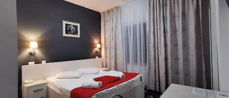 A hotel room with a bed, bedside tables, a lamp, a painting, and a window with curtains.