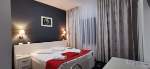 Una habitación de hotel con cama, mesitas de noche, lámpara, un cuadro y ventana con cortinas.