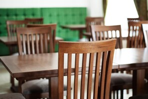 A dining area with wooden tables and chairs, a green upholstered wall, and a window with curtains.