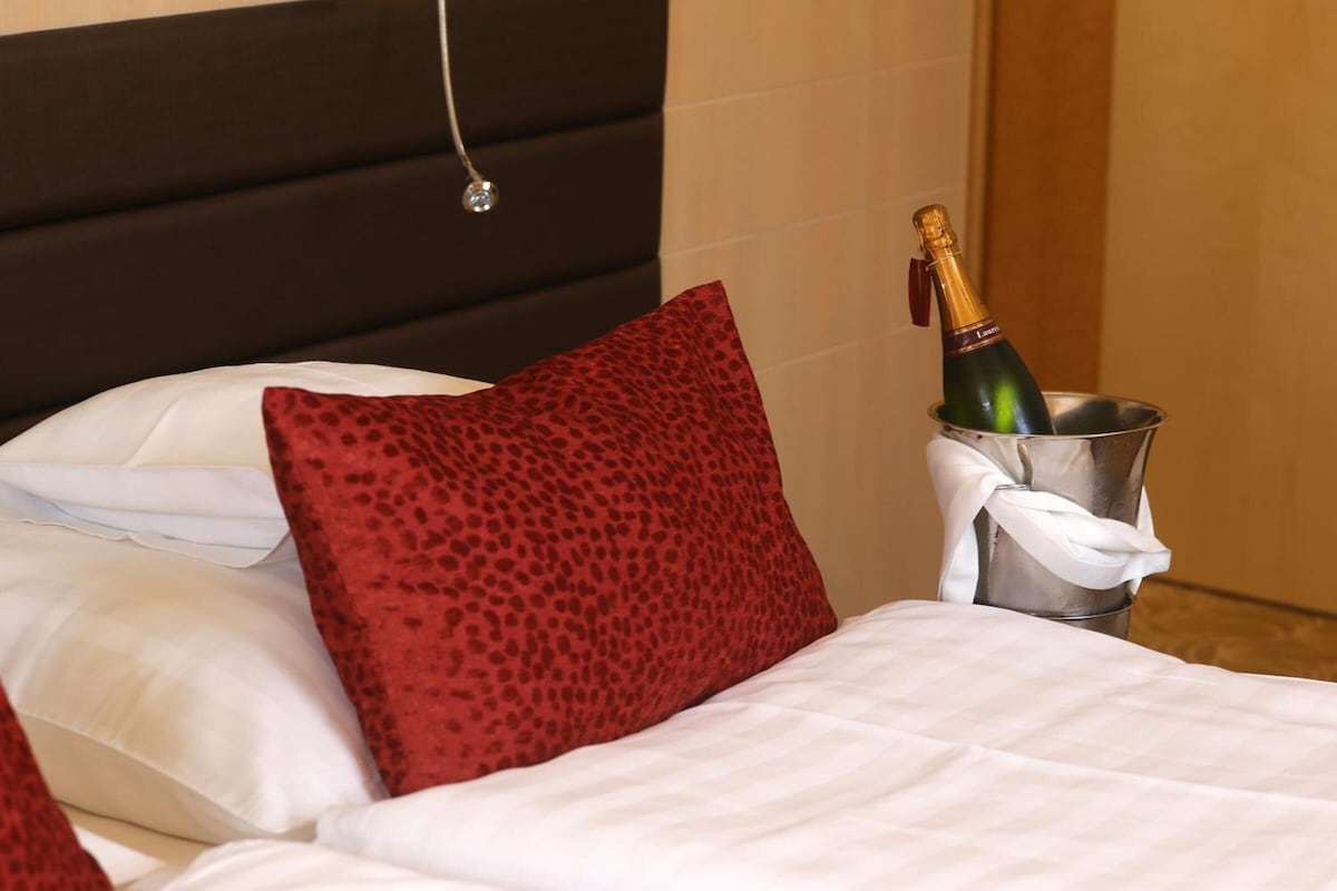 Una cama con almohadas blancas y rojas, una botella de champán en un cubo plateado y un cabecero oscuro.
